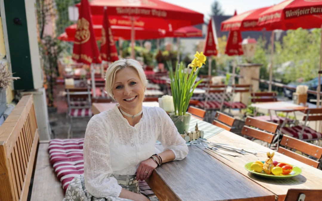Katrin, die Chefin vom Wurzn, lächelt im sonnigen Biergarten an einem Holztisch mit Frühlingsblumen und roten Sonnenschirmen im Hintergrund.